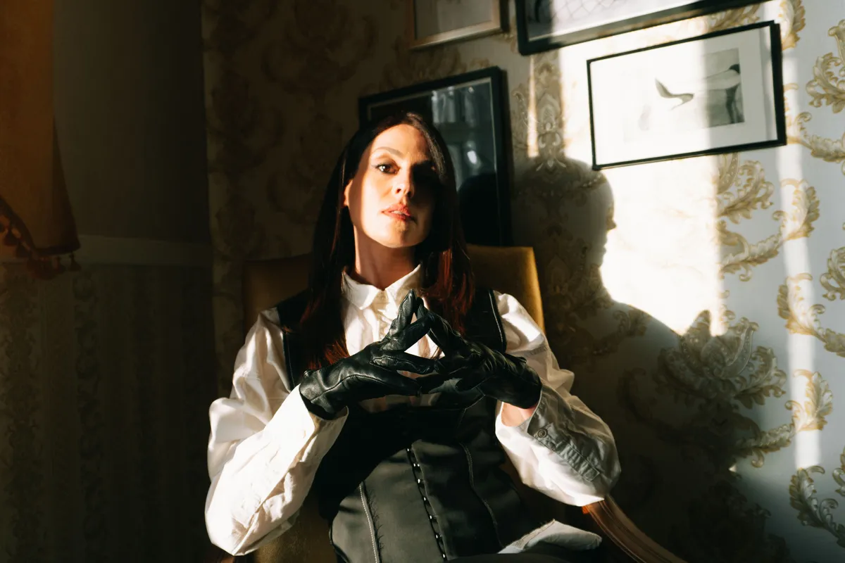 Aido Akida, femdom based in Berlin. She is wearing a white shirt and clack corset paired with black leather gloves. She is bathed in dramatic light.