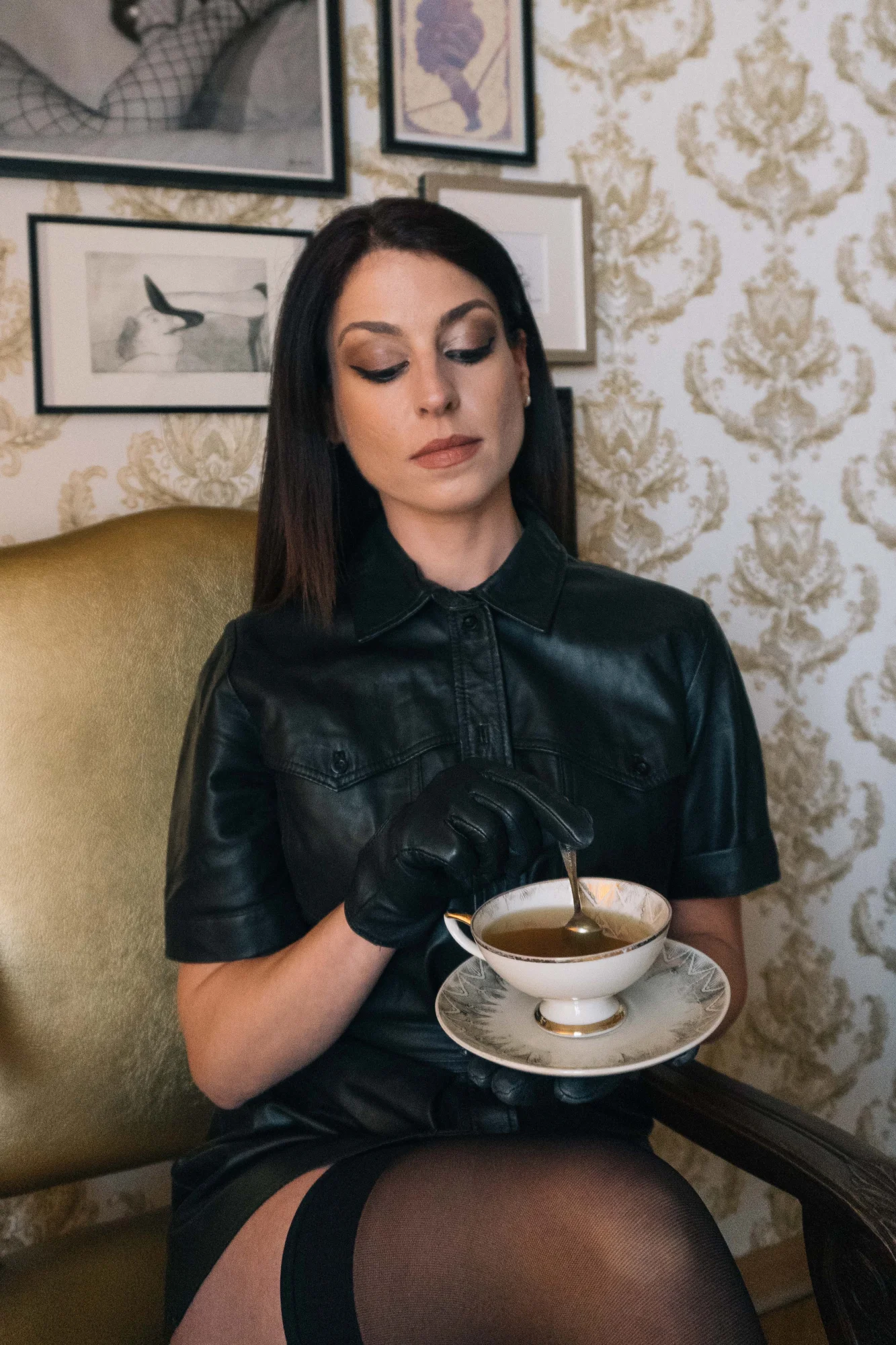 Aido Akia, dominatrix based in Berlin, she is wearing a black leather dree, black gloves, she is holding a cup of tea.
