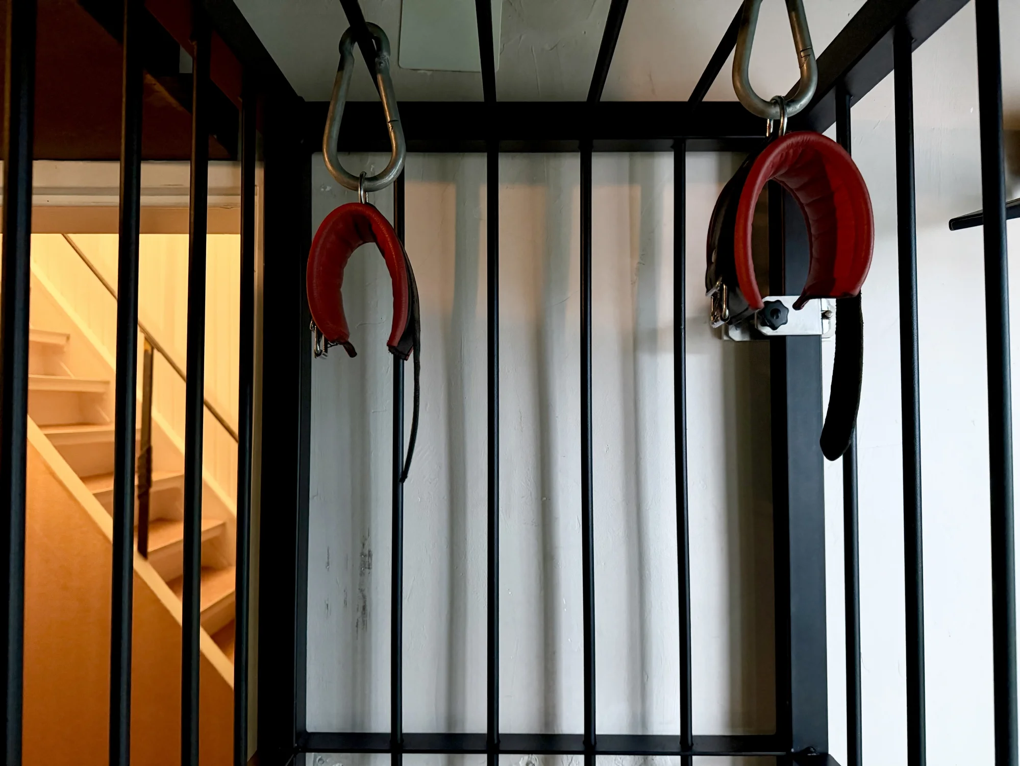 A picture of a black cage and a pair of leather handcuffs hanging from its ceiling. Stairs and orange lights as a background