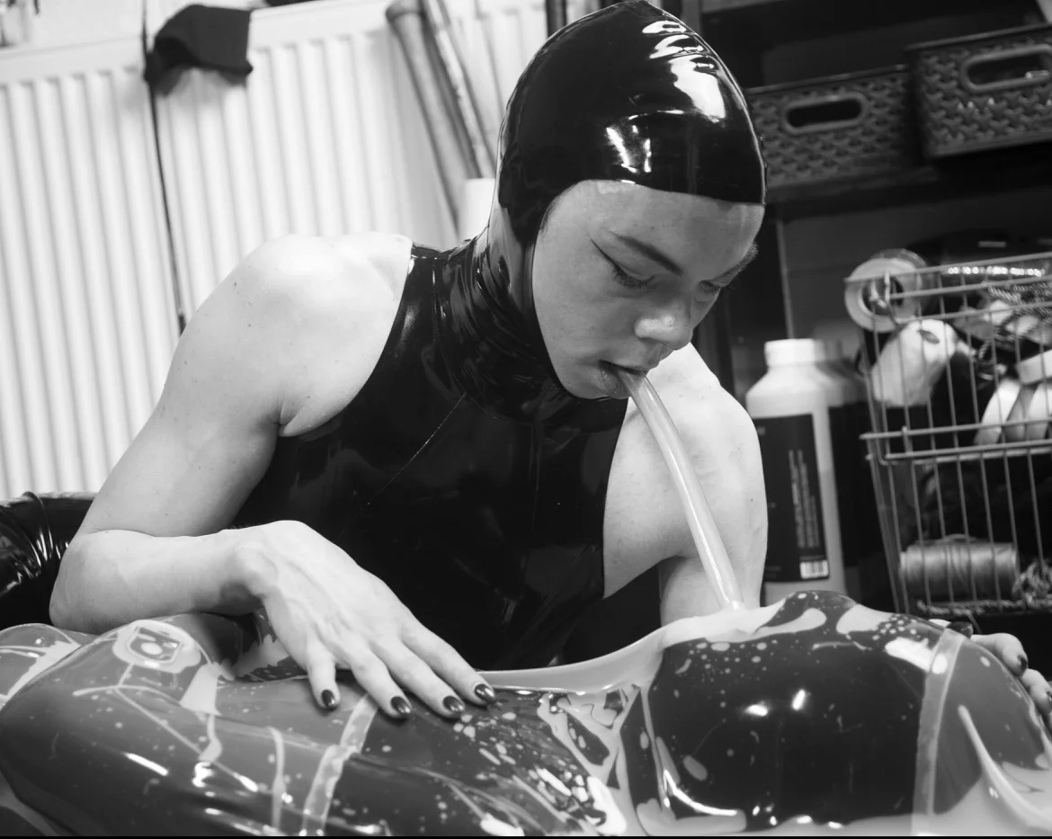 Woman wearing a black latex hood giving air to a men inside a vacuum bed through a PVC tube.