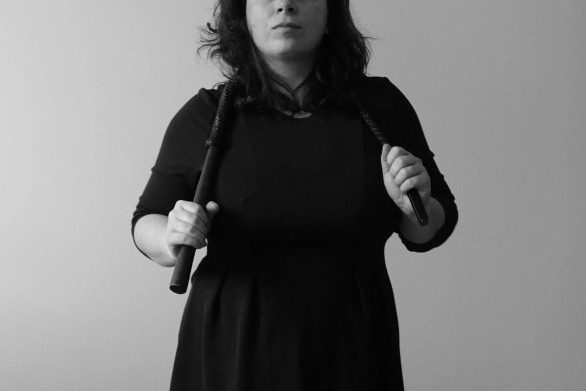 Architect holds a whip around her neck wearing a black dress and faces the camera sternly.