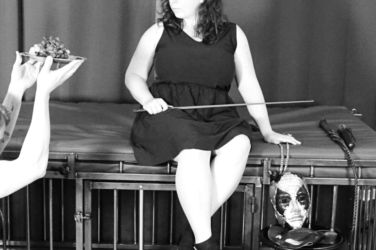 Architect is seated, holds a cane, wearing a black dress and faces a sub offering her grapes on a tray.