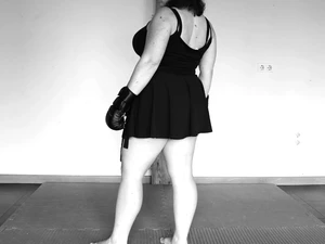 The Architect is standing on a mat with boxing gloves and a short black dress, with her back to the camera.