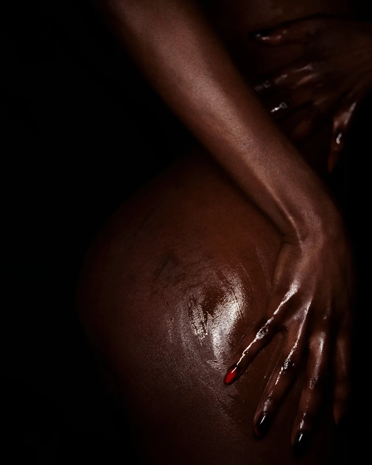 A black skinned person laying on their side. One hand on resting on the hip, with red and black nail polish. While the otherhand crosses her belly, reaching the upper part of the other arm.
