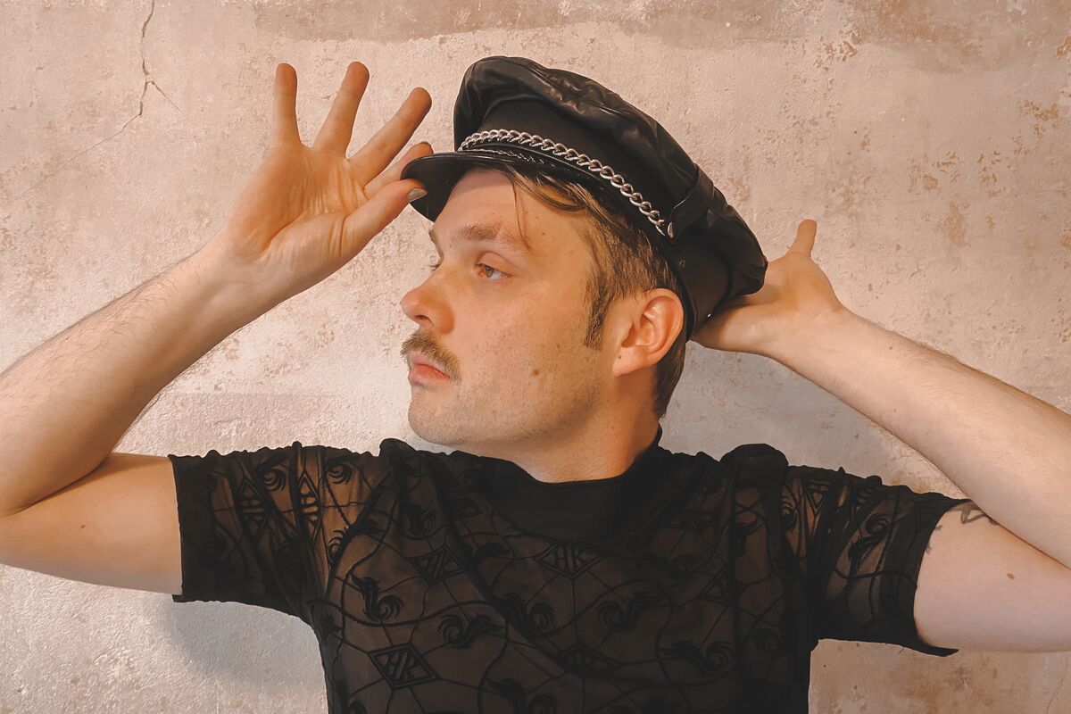 Dom Ferdinand Krista in einem schwarzen, leicht durchsichtigen Mesh-Top mit einer Mütze aus Leder, die er sich elegant zurechtschiebt