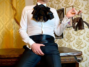 Ferdinand Krist steht an einen alten Schreibtisch gelent, er trägt ein weißes Hemd und eine schwarze Hose, einen großen Kragen aus schwarzem Latex, in einer Hand hält er eine Ledermanschette, mit d4er anderen greift er in seinen Schritt