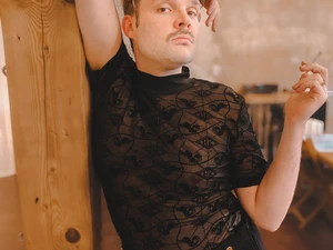Ferdinand Krista steht an einen Holzpfosten im Kursraum des Studio LUX gelehnt in einem schwarzen Mesh Top und schwarzer Hose, einen Arm über den Kopf gelegt, den anderen angewinkelt in der Hüfte mit Zigarette in der Hand