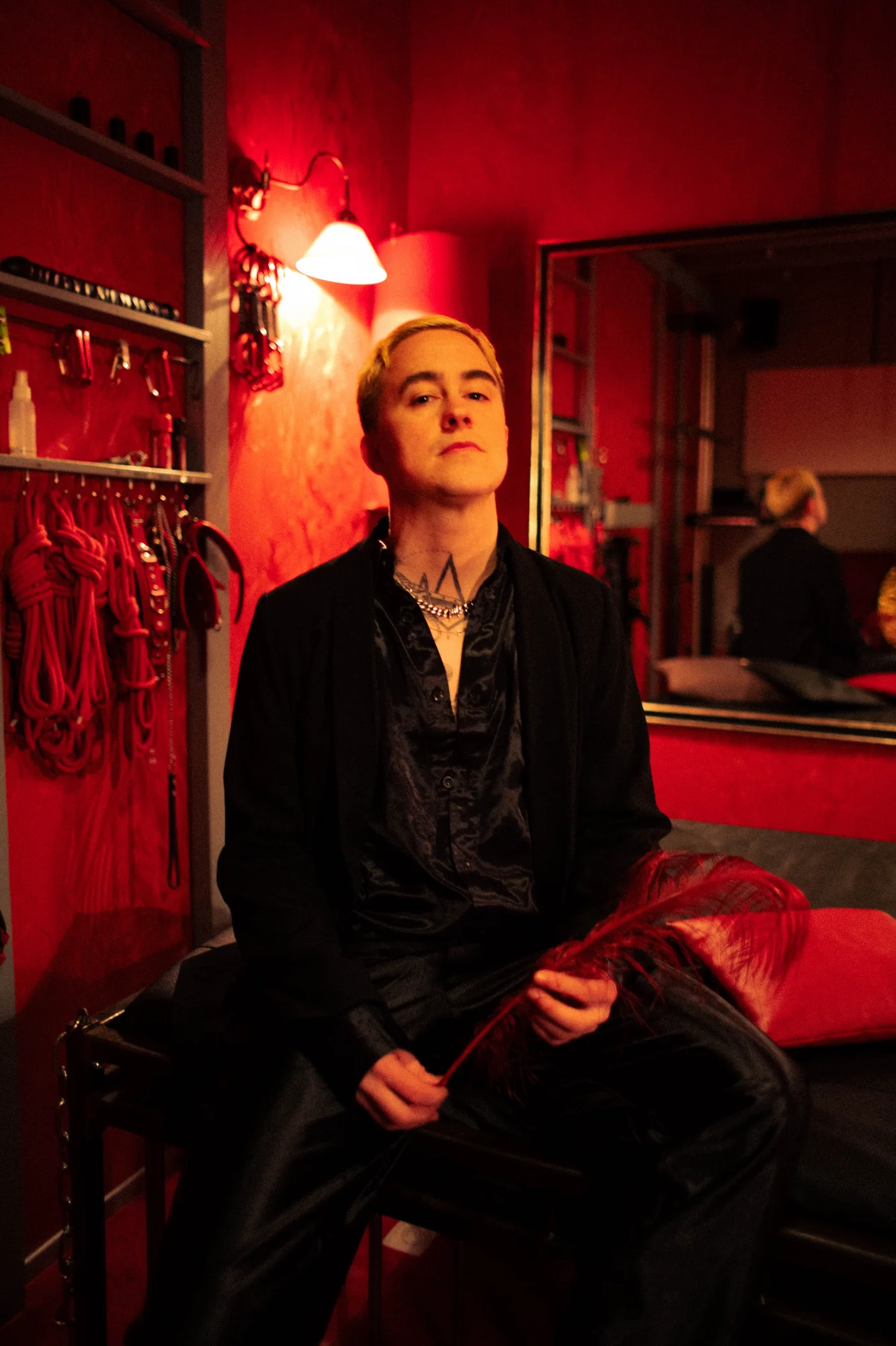 White tattooed person with short hair in a black suit sits on a leather couch holding a red feather; mirrors and hanging accessories can be seen in the background.