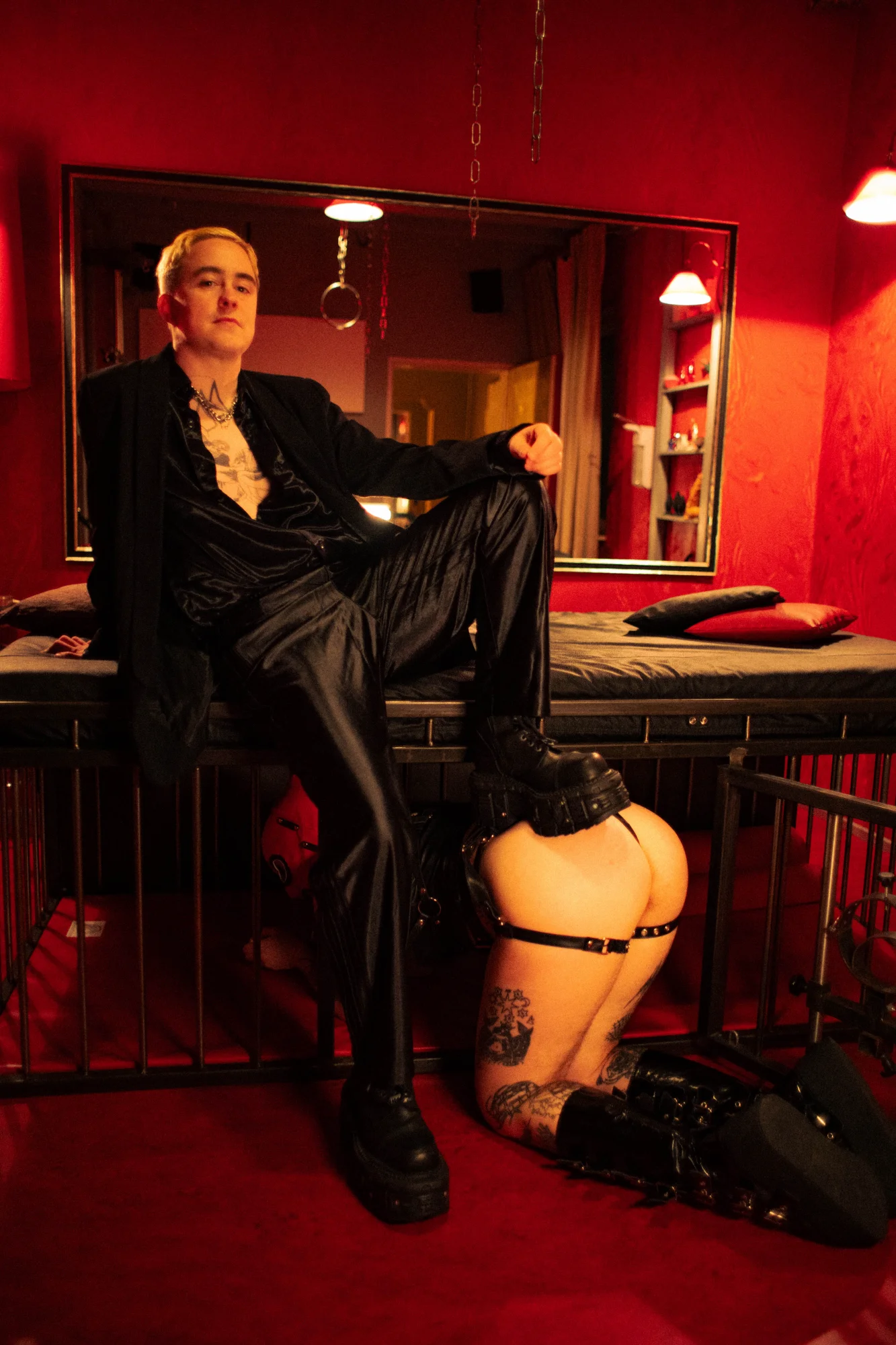 White tattooed person with short hair, a half-unbuttoned shirt, and a black suit sits with one leg raised on a leather couch; another person kneels in front of them wearing black leather clothing; the room is lit in red.