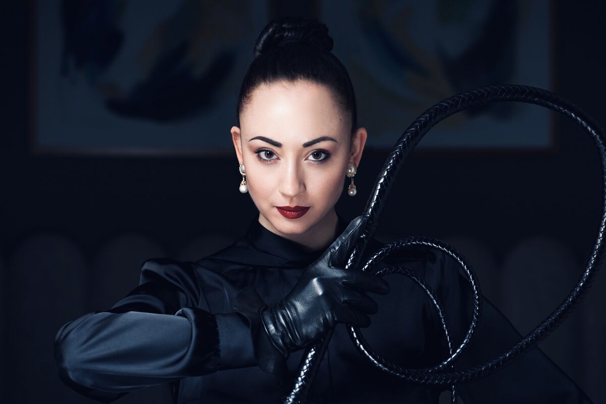 Lady Mephista holding her whip. Lady Mephista looks directly at you. She wears a pair of leather gloves and a silk blouse paired with pearl earrings. She wears her hair in a tight knot. She holds her whip with her right arm in front of her.