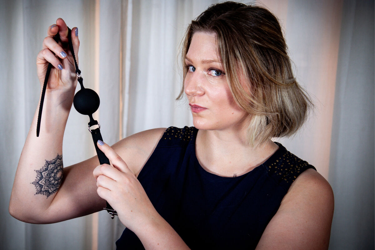 Bizarrlady Lina Twist holds out a black ball gag to you at Studio LUX Berlin.
