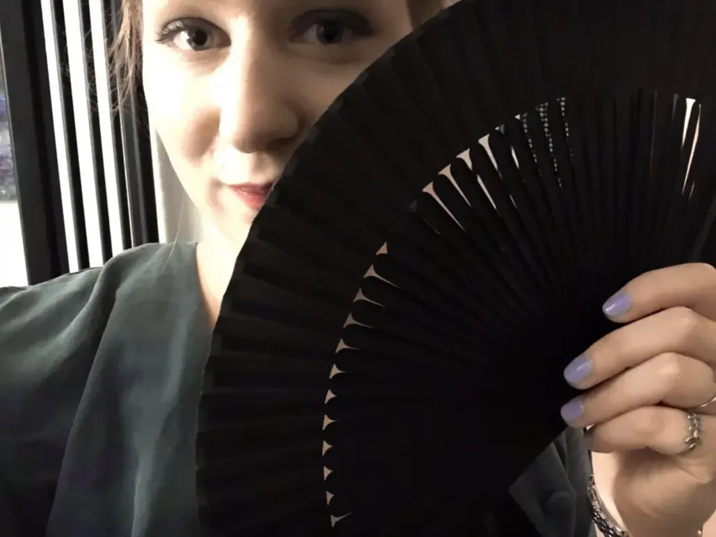 Lina Twist lächelt dich verschmitzt an, ihr Gesicht ist halb von einem schwarzen Fächer verdeckt.