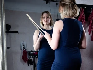 Domina Lina Twist steht mit Rohrstock in der Hand vor dem Spiegel und lächelt mit Vorfreude im Studio LUX Berlin.