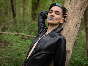Luna lehnt an einem Baum, hat den Arm hinter dem Kopf, trägt blauen Lippenstift und schaut in die Kamera, der schwarze Latexcatsuit ist leicht geöffnet, im Hintergrund ist es grün