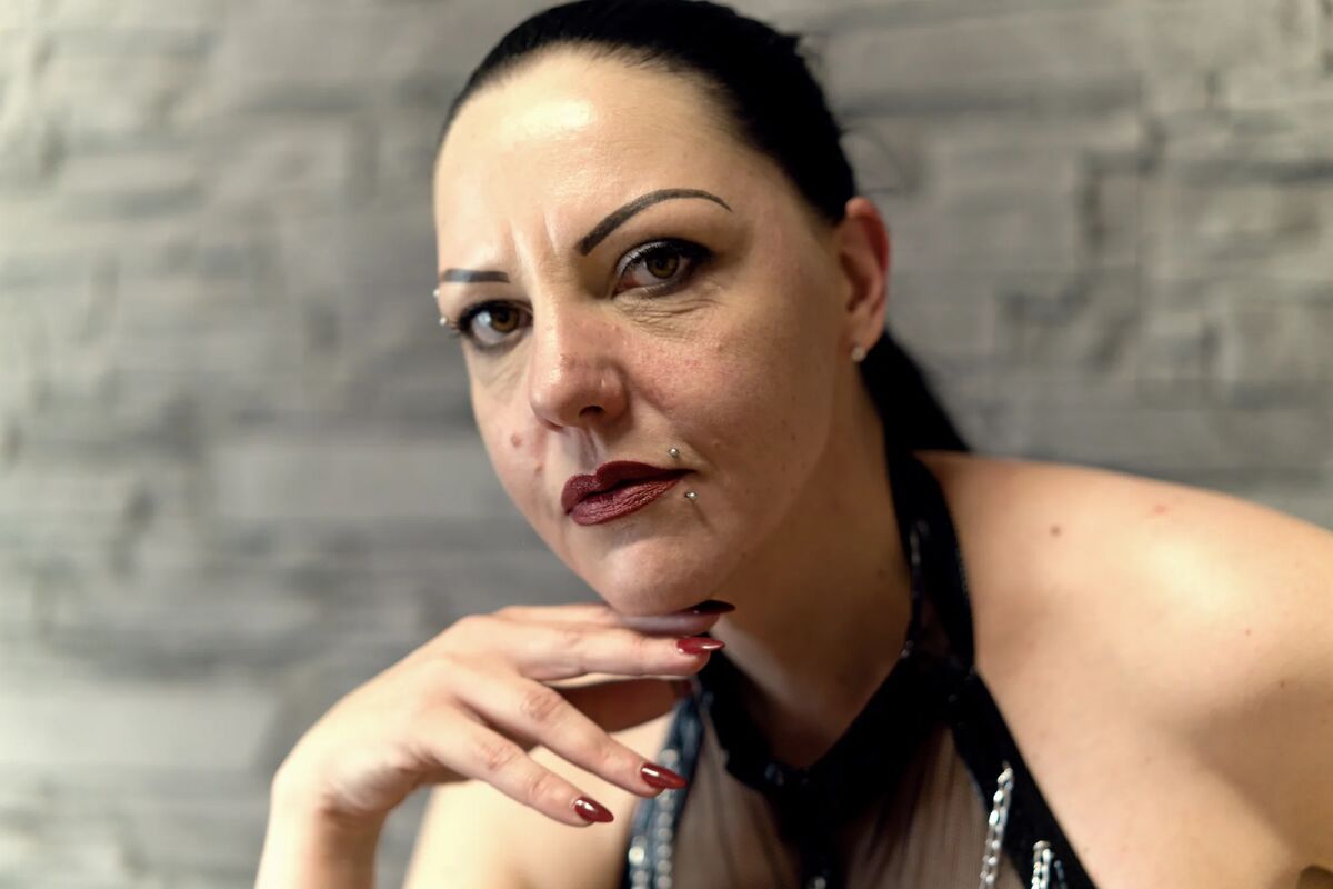 Nahaufnahme-Porträt einer Frau mit dunklem, zurückgebundenem Haar, dunklem Lippenstift und Piercings. Sie blickt direkt in die Kamera, während ihre Hand mit rot lackierten Nägeln an ihrem Kinn ruht.