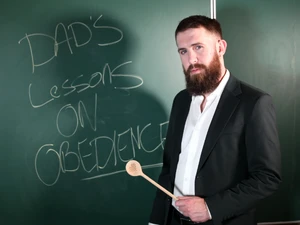 Bearded Man In Suit Holding Wooden Spoon In Front Of Chalk Board That Reads Dad's Lessons On Obedience