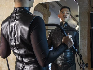 Ron trägt ein eng anliegendes schwarzes Lederoberteil und blickt in einen Wandspiegel, hält eine Peitsche in der Hand im BDSM Studio LUX