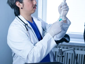 Ron mit weißem Kittel und Stethoskop zieht mit Handschuhen eine Spritze auf im BDSM Studio LUX