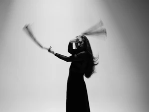 Ron tanzt barfuß auf leerer Bühne und schwingt zwei Peitschen, lange Haare und fließendes dunkles Outfit; dynamische Schwarzweißaufnahme.