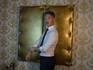 Ron steht im Anzug vor einer luxuriösen goldenen, gepolsterten Wand im BDSM Studio Lux
