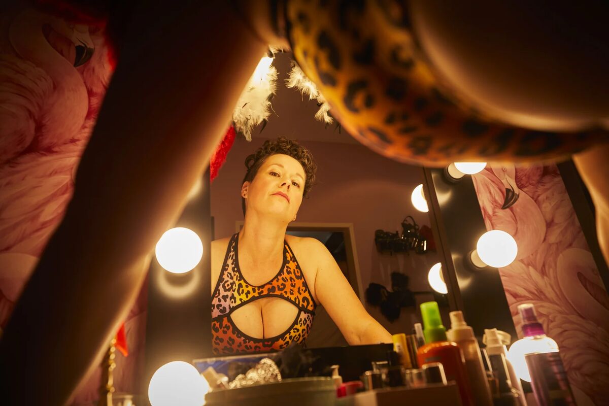 Bizarrlady Tamara Solidor in buntem hautengen Leo-Top mit tiefem Ausschnitt steht vorübergebeugt und schaut über den Spiegel auf dich herab in einer weichen angenehmen Lichtstimmung