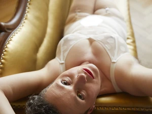 Tamara Solidor liegt in weißer Reizwäsche auf dem goldenen Sofa im Studi-Raum des Studio LUX und hält die Arme über den Kopf verschränkt und schaut auffordernd in die Kamera