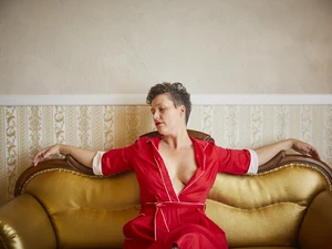 Tamara Solidor sitzt in einem roten Kleid mit tiefem Ausschnitt auf einem goldenen Sofa, sie hat die Arme ausgebreitet auf der hinteren Lehne ruhend und schaut zur Seite