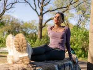Violet Beau sits on a wall with her legs outstretched infront of her, she is making a cheeky expression an looking off into the distance. The setting is a lovely sunny, green park